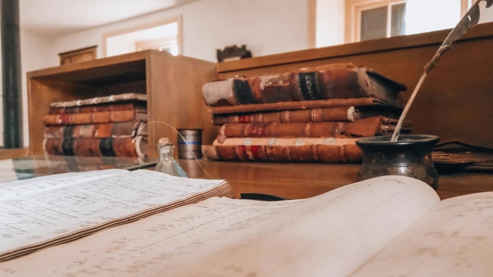 Handwritten ledgers open on a desk beside a quill and ink pot, with bound volumes behind
